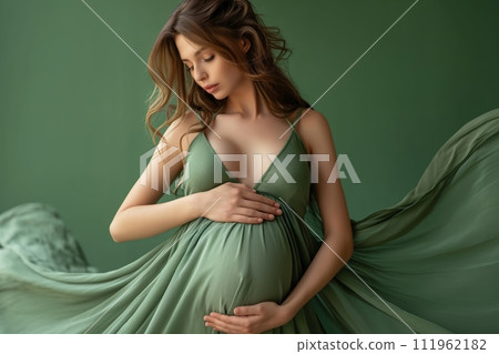 9 month pregnant woman wears fashionable green dress touching belly against green background 111962182