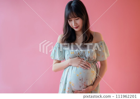 9 month pregnant Asian woman wears flower printed dress touching her belly against pink background 111962201