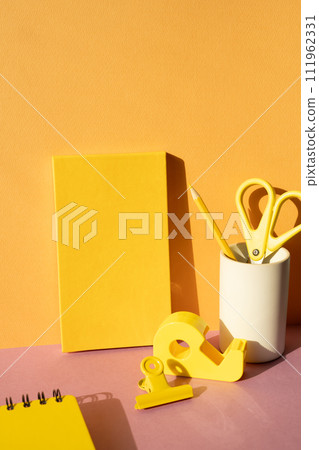 Office supply stationery. Yellow notebook, colored pencil, scissors, tape dispenser, clip on pink desk. orange wall background. workspace 111962331