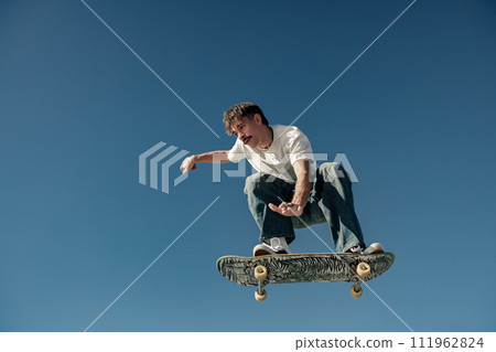 Active man doing tricks in the air on his skateboard at the skate park on blue sky background 111962824