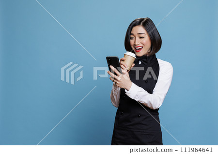 Cheerful asian waitress laughing while drinking takeaway coffee and scrolling social media on smartphone. Smiling woman receptionist enjoying beverage to go and using mobile phone Cheerful asian waitress laughing while drinking takeaway coffee and scrolling social media on smartphone. Smiling woman receptionist enjoying beverage to go and using mobile phone 111964641