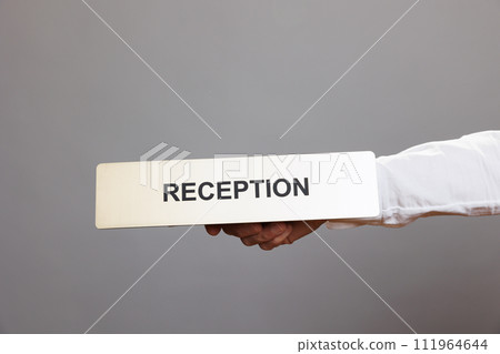 Hotel administrator hand holding reception stainless steel plate closeup. Hospitality service receptionist arm showing tabletop metal name sign close view in studio on gray background 111964644