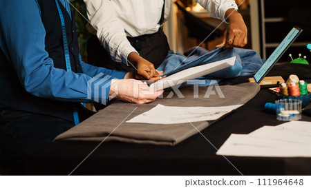 Old craftsman and trainee working in atelier, checking sketches of clothes to manufacture fashion design. Beginner learning the art of handcraft from experienced tailor. Handheld shot. Old craftsman and trainee working in atelier, checking sketches of clothes to manufacture fashion design. Beginner learning the art of handcraft from experienced tailor. Handheld shot. 111964648