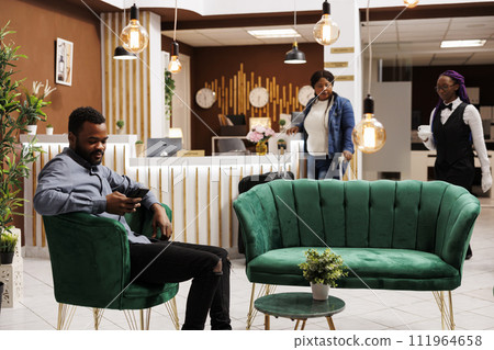 African American millennial guy looking at smartphone screen reading text message while relaxing in lounge area at luxury resort, man traveler holding phone using hotel wifi while sitting in lobby 111964658