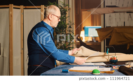 Male business owner making clothes, looking at sketches in tailoring workshop to create custom made items. Senior tailor working with tools and fabric to make modern products. Handheld shot. Male business owner making clothes, looking at sketches in tailoring workshop to create custom made items. Senior tailor working with tools and fabric to make modern products. Handheld shot. 111964721