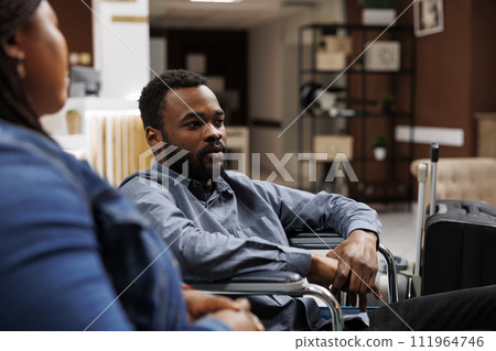 African American man tourist with physical impairment in wheelchair waiting with luggage in hotel lobby, arriving at rehabilitation center for disabled. Inclusive accessible tourism 111964746