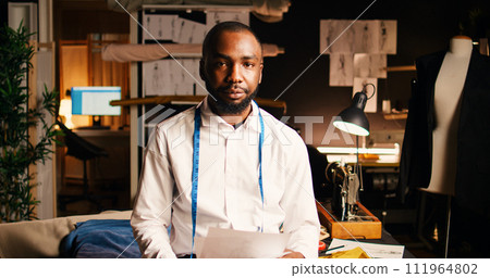 Couturier checking sketches and designs, having tailoring meter around his neck. African american tailor reviewing drawings outlines to manufacture fashion items, needlework concept. 111964802