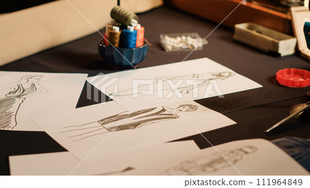 Tailoring sketches to design clothes on atelier workstation, drawings on papers with clothing line for new fashion collection. Designer atelier with industrial tools. Close up. Tailoring sketches to design clothes on atelier workstation, drawings on papers with clothing line for new fashion collection. Designer atelier with industrial tools. Close up. 111964849