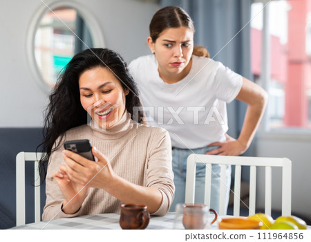 Woman using smartphone at home, her angry friend standing behind and watching Woman using smartphone at home, her angry friend standing behind and watching 111964856