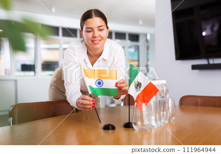 A woman in a white blouse with friendly smile arrangs flags of Mexico and Iran for meeting. 111964944