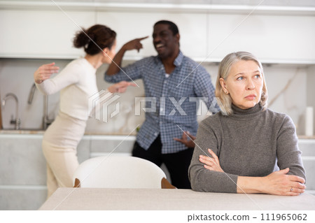 Old woman sitting at the kitchen table with her back to couple quarreling together Old woman sitting at the kitchen table with her back to couple quarreling together 111965062