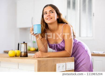 Portrait of girl in purple nightgown holding cup of coffee in her hands at home kitchen 111965204