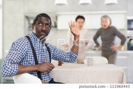 Tense man listens to two women shouting at him in kitchen Tense man listens to two women shouting at him in kitchen 111968443