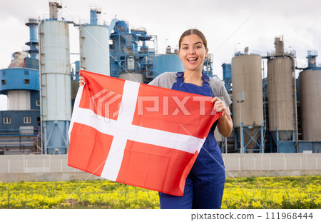 Happy young woman worker with flag of Denmark against background of factory Happy young woman worker with flag of Denmark against background of factory 111968444