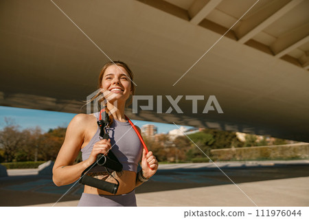 Athletic woman with a resistance band slung around her neck standing outdoors and looks away 111976044
