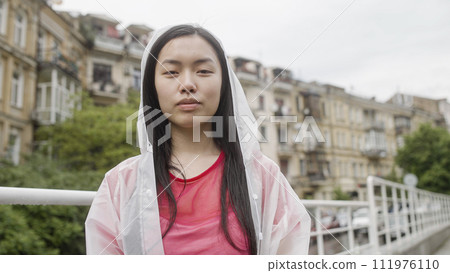 Portrait of young Asian woman in raincoat standing on the street, city life 111976110