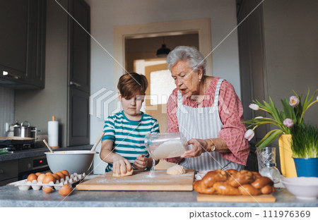 Grandmother with grandson preparing traditional easter meals, kneading dough for easter cross buns. Passing down family recipes, custom and stories. 111976369