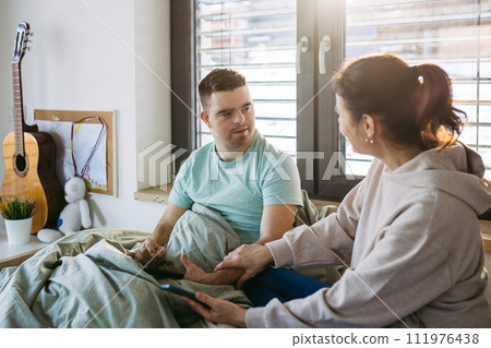 Young man with down syndrome sitting in bed with mom, looking at smartphone in morning. Morning routine for man with disability. Young man with down syndrome sitting in bed with mom, looking at smartphone in morning. Morning routine for man with disability. 111976438
