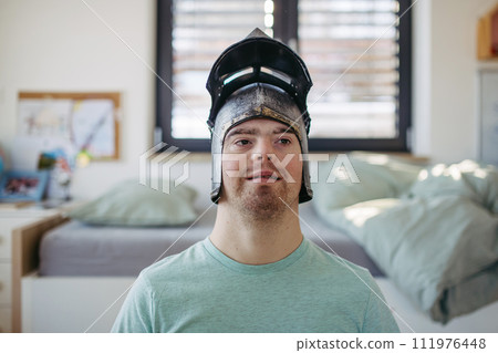 Portrait of smiling man with down syndrome at home with knight's helmet on head. Portrait of smiling man with down syndrome at home with knight's helmet on head. 111976448