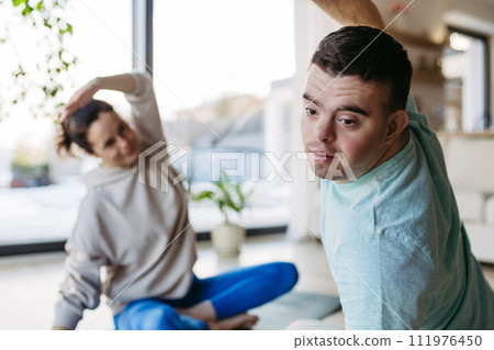 Young man with Down syndrome exercising at home with mother. Workout routine for disabled man. 111976450
