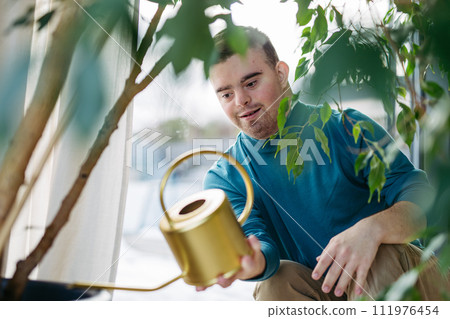 Young man with Down syndrome taking care of indoor plants, watering them, looking at camera through handle of a metal watering can. Young man with Down syndrome taking care of indoor plants, watering them, looking at camera through handle of a metal watering can. 111976454