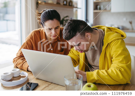 Mom teaching young man with down syndrome, using laptop. Telehealth consultation with doctor. Mom teaching young man with down syndrome, using laptop. Telehealth consultation with doctor. 111976459