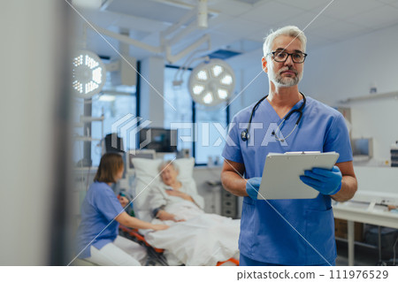 Portrait of handsome male doctor, patient in hospital bed behind. ER doctor examining senior patient, reading her medical test, lab results in clipboard. 111976529