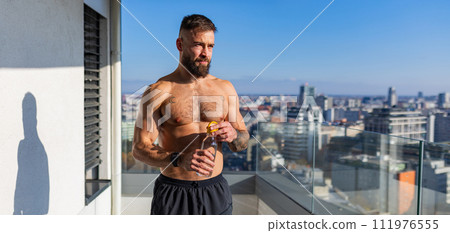 Handsome man with bare chest standing on balcony and enjoying beautiful view on city. Concept of selfcare and enjoying life for young people. Handsome man with bare chest standing on balcony and enjoying beautiful view on city. Concept of selfcare and enjoying life for young people. 111976555