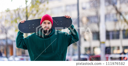 Stylish skateboarder with skateboard behind head, looking at camera. Young man skating outdoors in the city. Concept of skateboarding as sport and lifestyle. Stylish skateboarder with skateboard behind head, looking at camera. Young man skating outdoors in the city. Concept of skateboarding as sport and lifestyle. 111976582