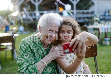 Young girl taking selfie with elderly grandmother at garden party. Love and closeness between grandparent and grandchild. 111976688