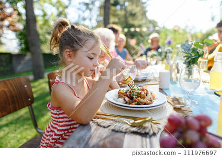 Portrait of a beautiful little girl sitting at table eating grilled food outdoors. Girl at family garden party. Portrait of a beautiful little girl sitting at table eating grilled food outdoors. Girl at family garden party. 111976697
