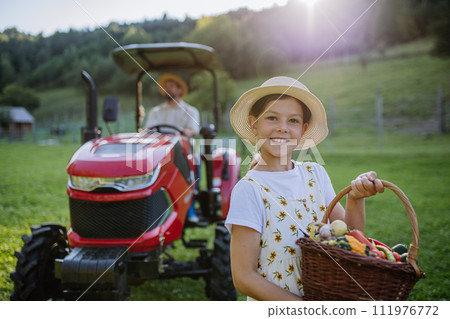 Girl on farm holding harvest basket full of fresh vegetables. Harvesting crops, collecting vegetables on family farm. Concept of multigenerational farming. 111976772