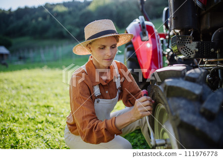 Female farmer fixing a wheel on a tractor. Progressive farmer working on tractor on her own farm. Women in agriculture. Female farmer fixing a wheel on a tractor. Progressive farmer working on tractor on her own farm. Women in agriculture. 111976778