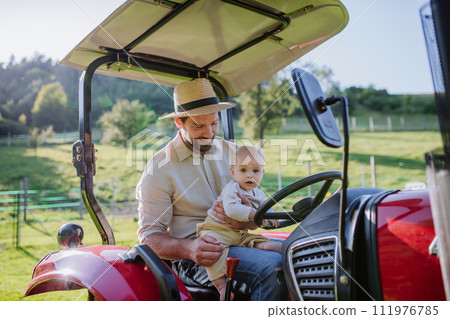 Farmer father riding tractor with his little baby son. Baby growing up on family farm. Concept of multigenerational farming. 111976785