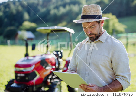 Farmer reading document in front of tractor on field. Grants, loans, funding opportunities for farmers. Farmer reading document in front of tractor on field. Grants, loans, funding opportunities for farmers. 111976789