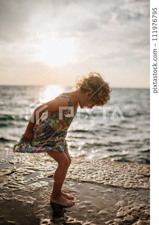 Young girl jumping in puddle on promenade by sea on sand beach. Happy girl in summer dress having fun. 111976795