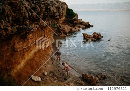 Brother helping sister seraching for shells in sand. Small girl in swimsuit playing at beach, crouching in water. 111976821