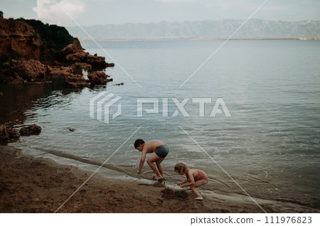 Brother helping sister seraching for shells in sand. Small girl in swimsuit playing at beach, crouching in water. Brother helping sister seraching for shells in sand. Small girl in swimsuit playing at beach, crouching in water. 111976823