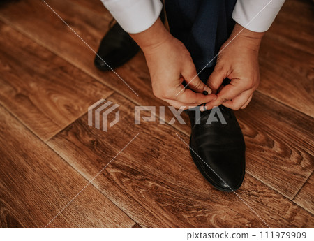 man in a white shirt and dark trousers is sitting on a chair tying the laces of his black boots 111979909