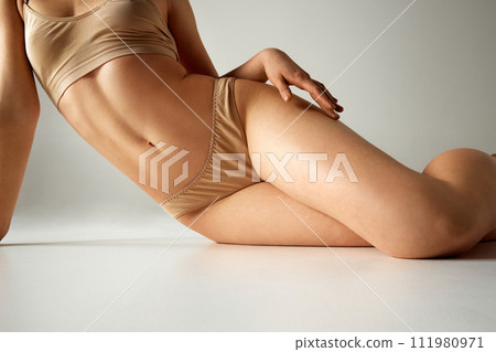 Cropped image of female body, slim waist, slender, smooth legs. Model posing in underwear against grey studio background. Cropped image of female body, slim waist, slender, smooth legs. Model posing in underwear against grey studio background. 111980971