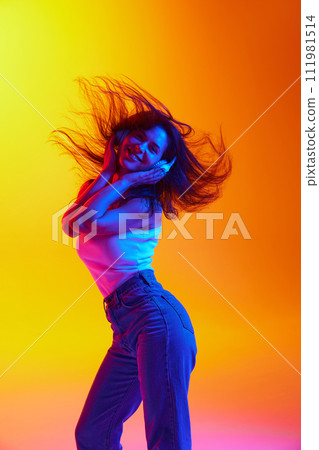 Woman with flowing hair in white top, jeans listening music against orange gradient background in neon light. Woman with flowing hair in white top, jeans listening music against orange gradient background in neon light. 111981514