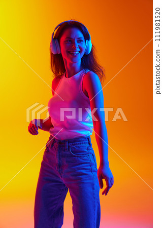 Portrait of young lady dressed casual outfit listening music in headphones against gradient studio background in neon light. Portrait of young lady dressed casual outfit listening music in headphones against gradient studio background in neon light. 111981520