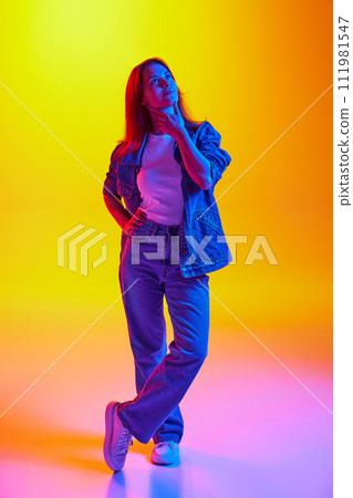 Full-length portrait of thoughtful, dreaming young lady dressed denim outfit against gradient orange-yellow background in mixed neon light. Full-length portrait of thoughtful, dreaming young lady dressed denim outfit against gradient orange-yellow background in mixed neon light. 111981547