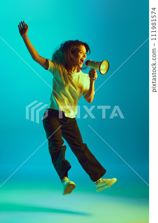 Full-length portrait young lady shouting important news to megaphone in yellow neon light against gradient studio background. Full-length portrait young lady shouting important news to megaphone in yellow neon light against gradient studio background. 111981574