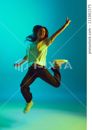 Full-length portrait of young beautiful woman jumping raising hands of joy in neon yellow lighting against gradient blue studio background. 111981575