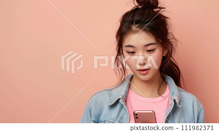 Pensive young Asian female in pink top and denim jacket looking at phone, isolated on beige. 111982371