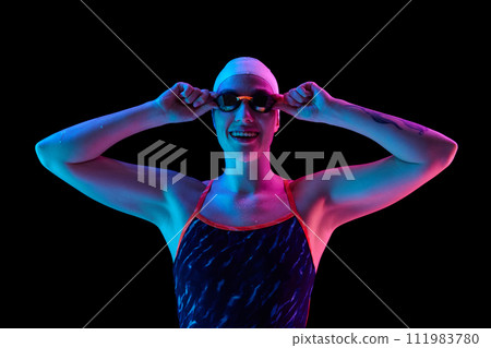 Portrait of positive, athletic woman in swimwear, goggles and cap looking at camera in vibrant neon light against black studio background. 111983780