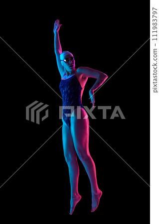 Dynamic portrait of young professional female swimmer poised in mid-action in vibrant neon light against black studio background. 111983797