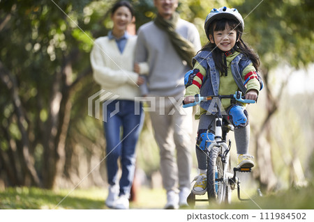 little asian girl riding bike in city park with parents in background little asian girl riding bike in city park with parents in background 111984502