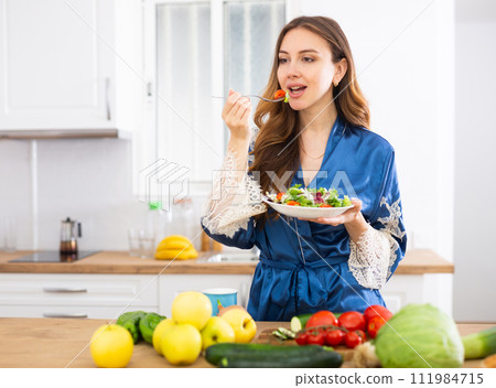 Young woman in blue silk robe enjoying vegetable salad in kitchen 111984715